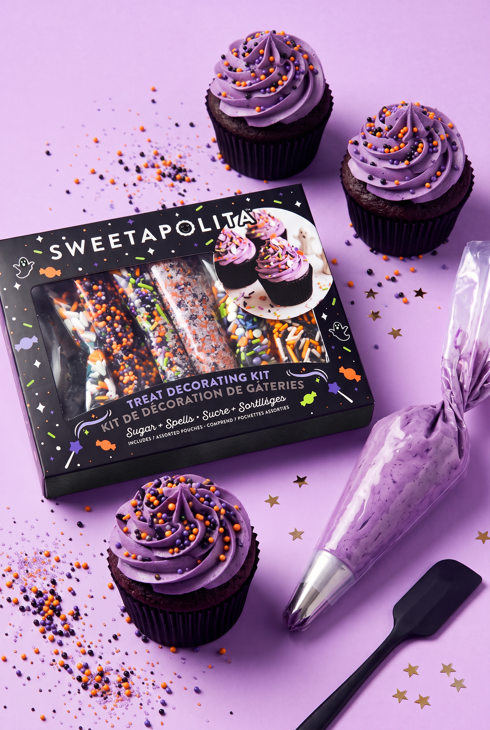 Sweetapolita Halloween treat decorating kit shown with a black gift box, a piping bag filled with lavender frosting, and three decorated cupcakes on a pastel purple background with sprinkles and gold stars.